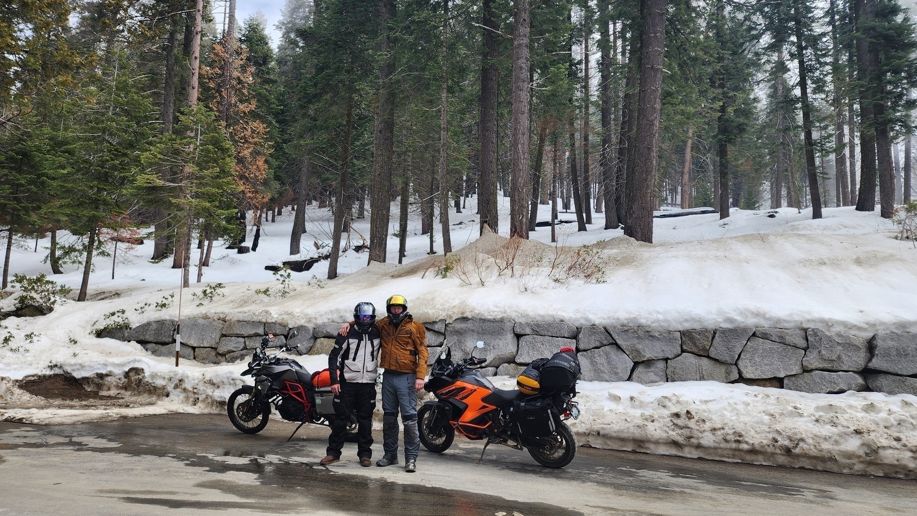 Two people wearing helmets stand next to two parked motorcycles on a snowy road surrounded by tall trees.