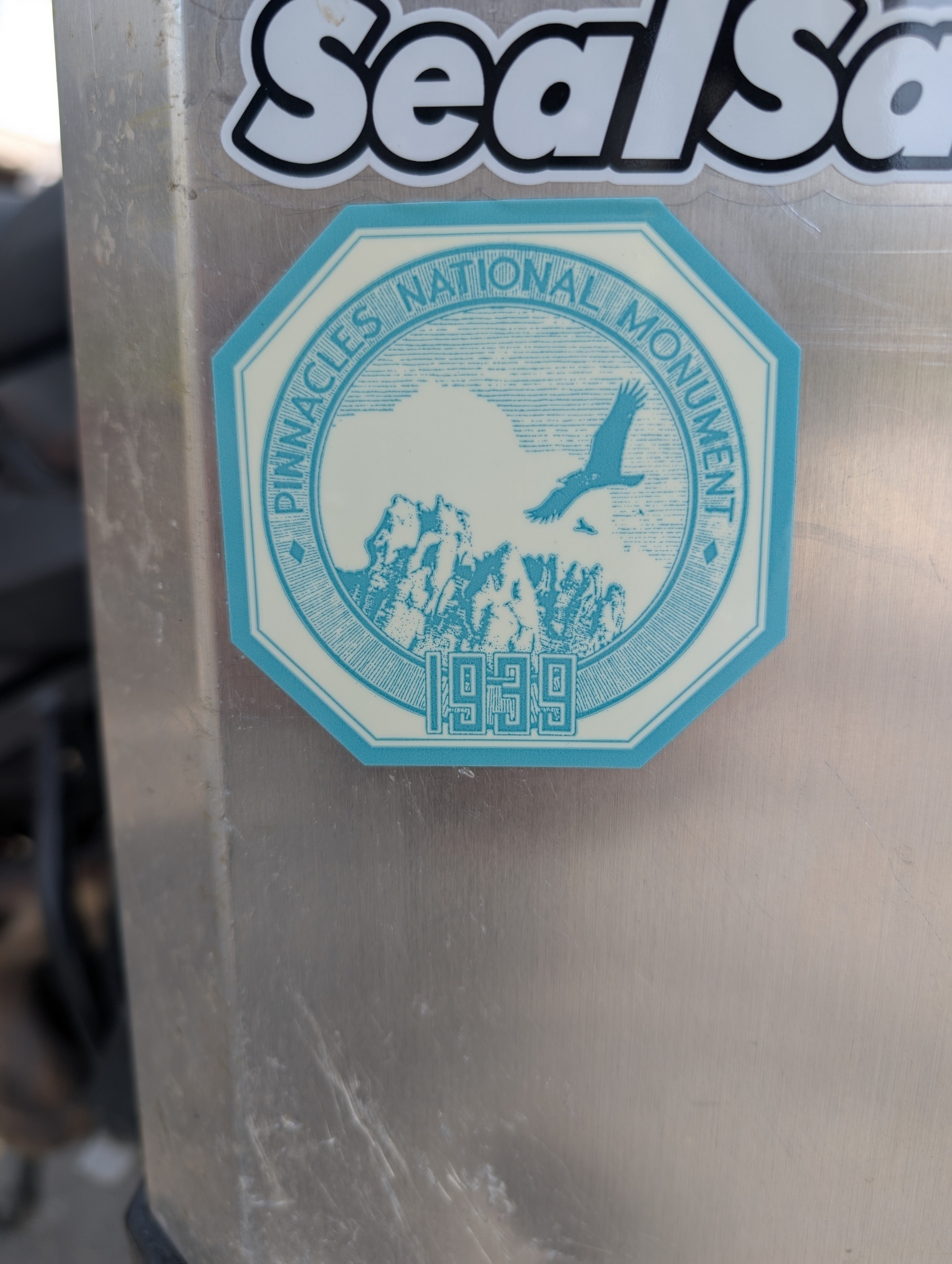 A reproduction of an antique window sticker depicts Pinnacles National Monument with a mountain, bird, and the year 1939. The sticker is affixed to the side of a motorcycle travel case.