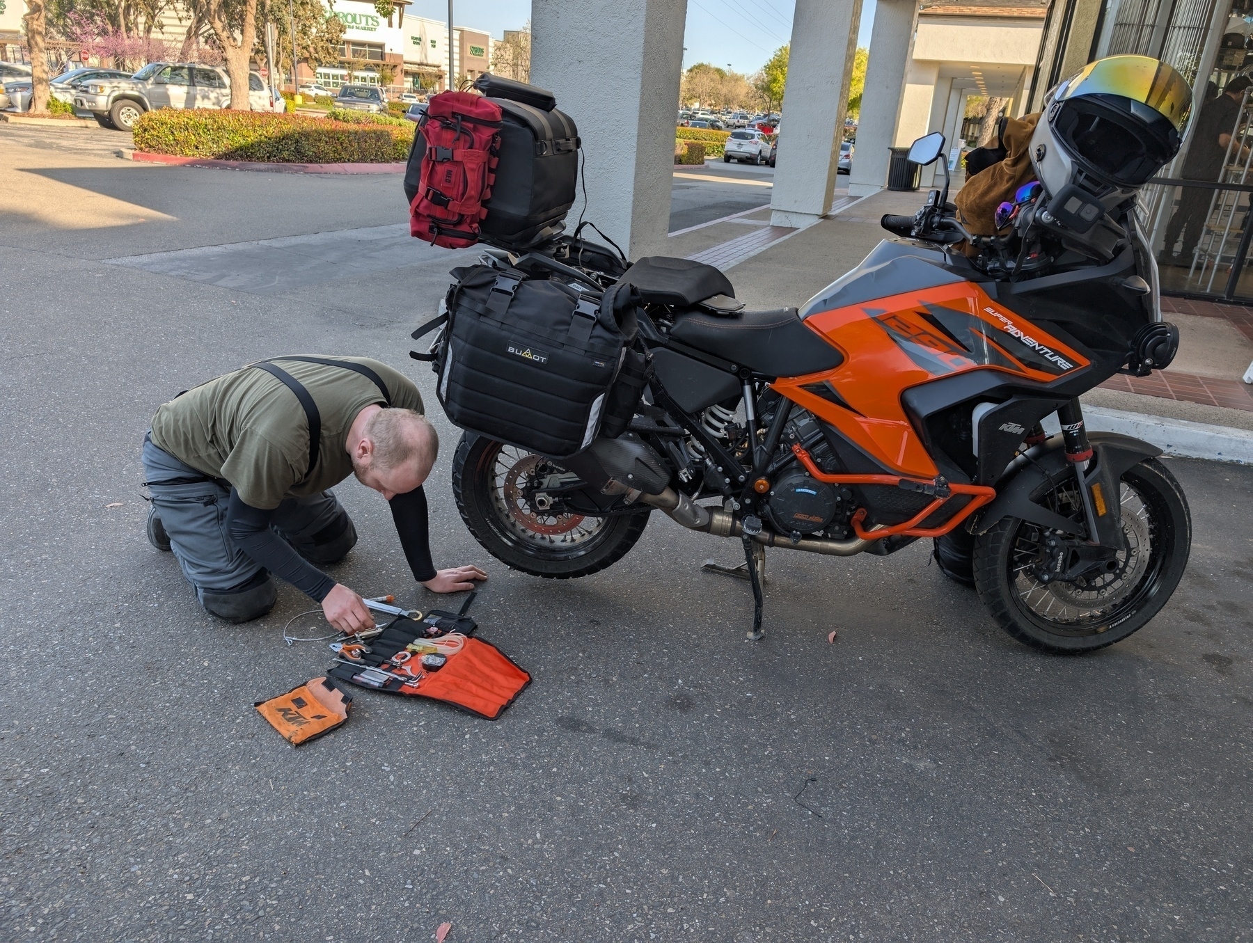 A person using a tool kit on the ground next to an orange and black KTM SuperAdventure motorcycle parked in a lot.