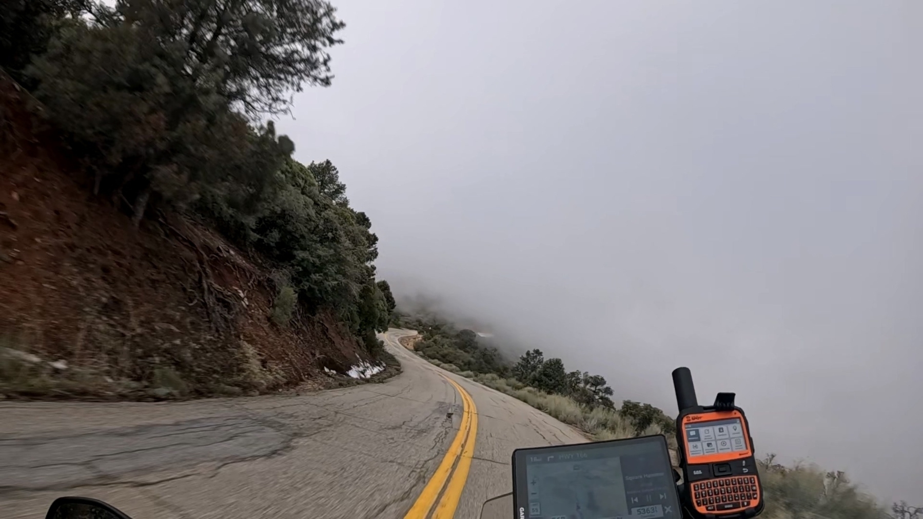 A view from the perspective of a motorcycle shows a GPS and dash in the foreground. In the background is Highway 166 in southern California. The road is narrow and winding through the mountains, with shrubs and some snow on the sides.