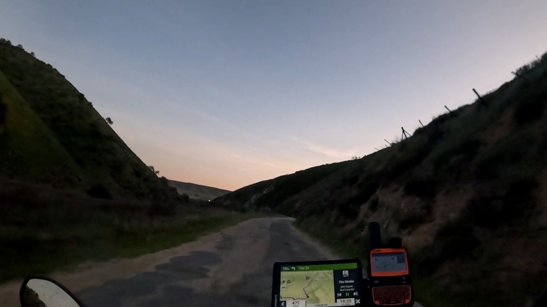 A narrow road winding through a valley at dusk, with a GPS device visible on a motorcycles handlebars.
