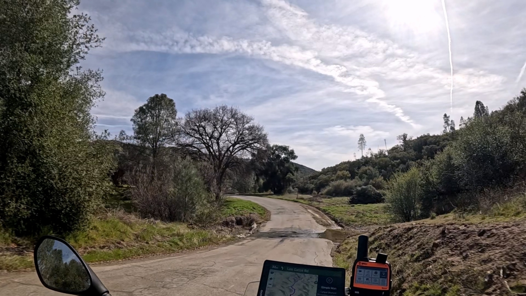 A winding road in a rural landscape is surrounded by trees and a small stream crossing the road.