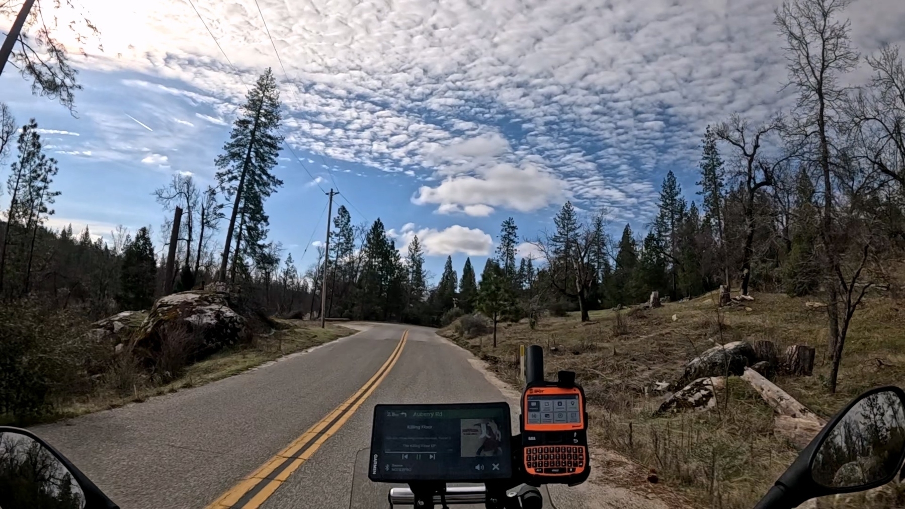 A winding road lined with trees and rocks leads through a scenic, forested area under a sky filled with scattered clouds.