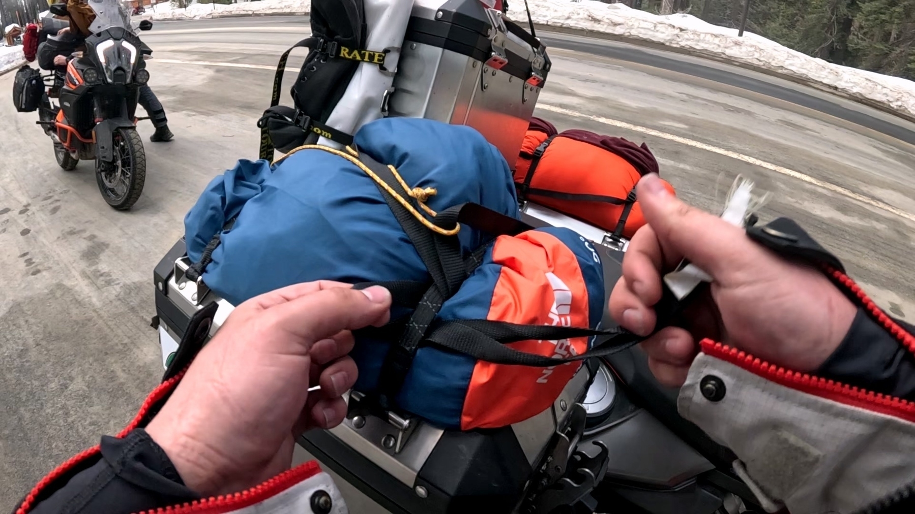 A person is adjusting a strap on a motorcycle loaded with camping gear and bags, while another motorcycle is visible in the background on a snowy roadside.