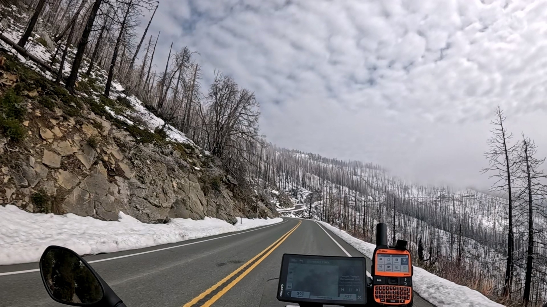 An empty road winds through a snowy, mountainous landscape with a cloudy sky overhead, flanked by rocky terrain and burned trees.