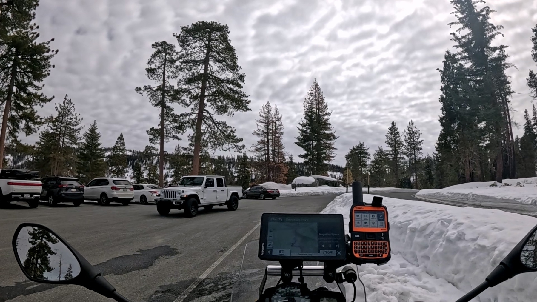 A snowy parking lot is bordered by trees, with several parked vehicles under a cloudy sky.