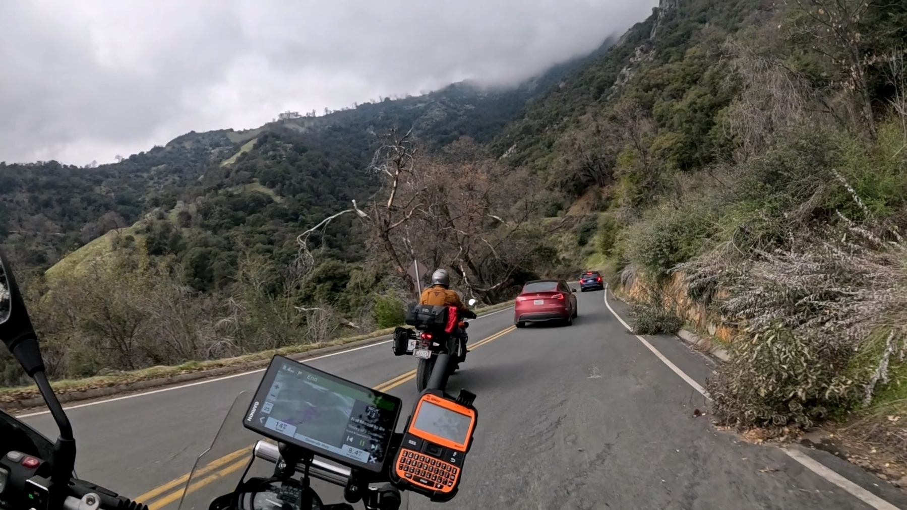 A motorcyclist follows cars on a winding road through a mountainous landscape.