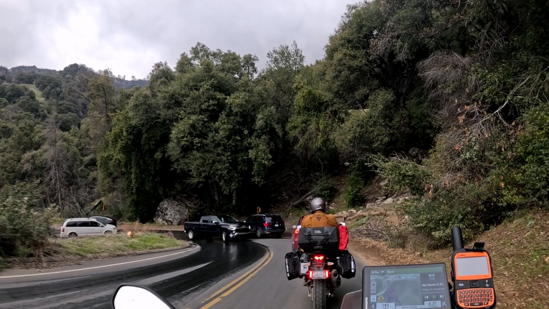 AA motorcyclist rides on a winding road surrounded by dense trees as a truck crosses over the center line, almost hitting the car in front.