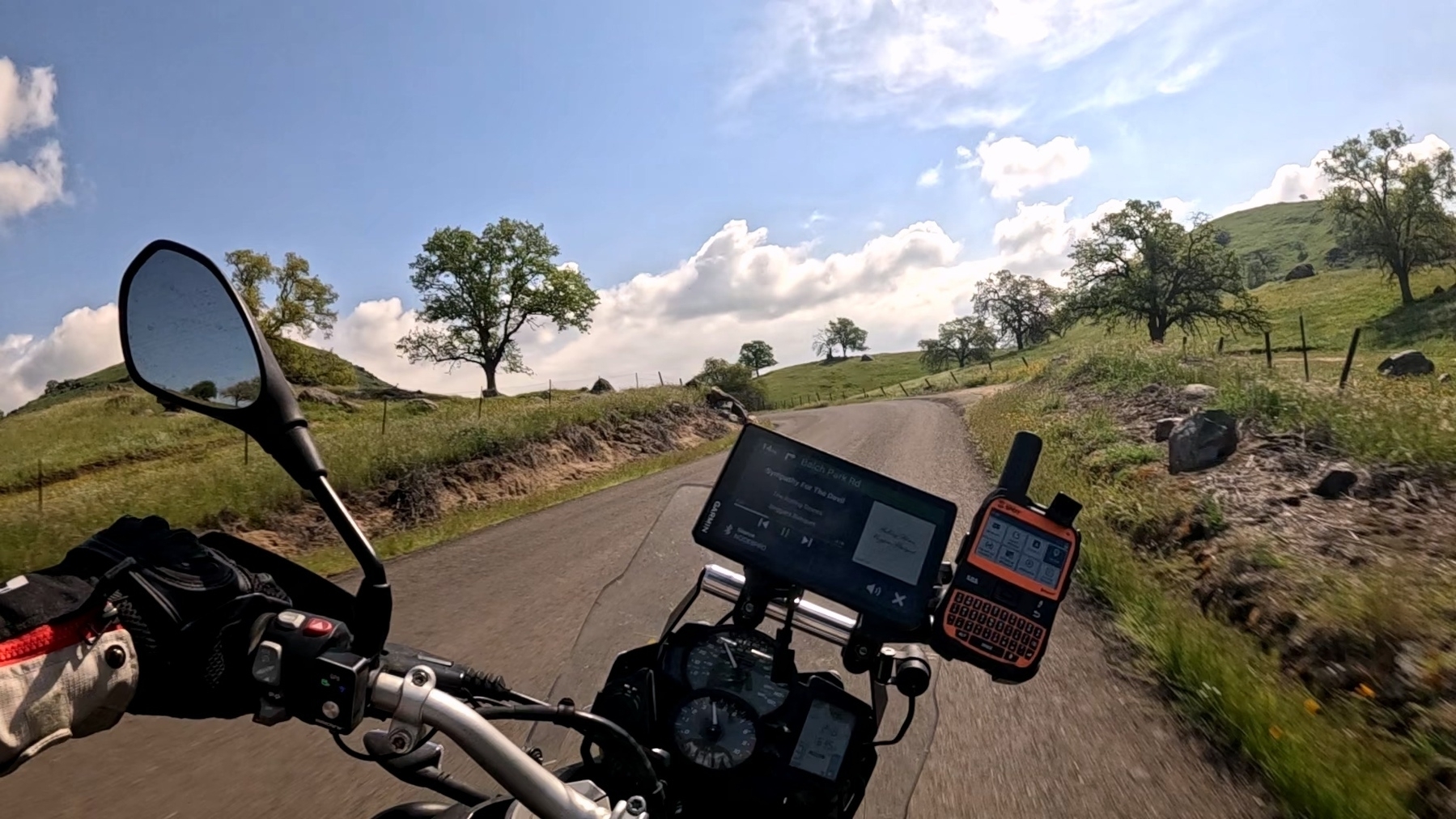 A motorcyclist rides through a scenic countryside road with hills and trees under a blue sky.