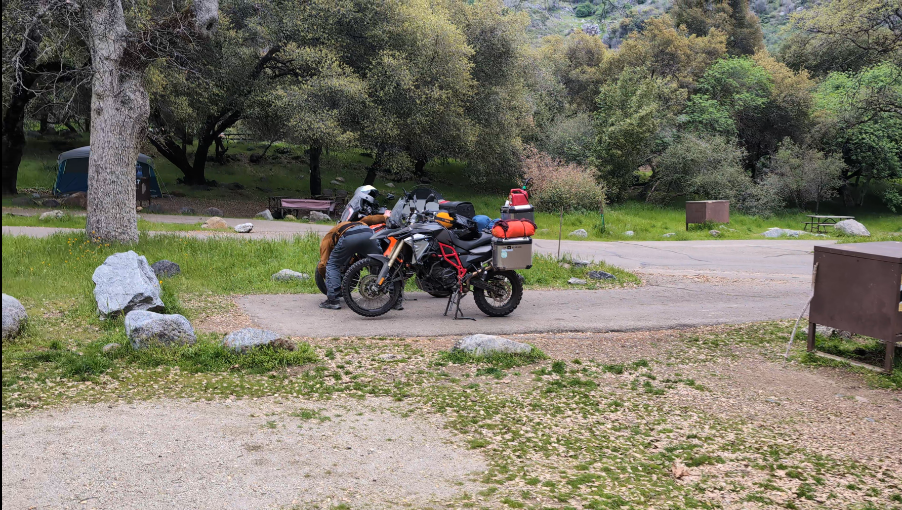 A person works on a motorcycle parked in a scenic, tree-filled campsite at potwisha campground